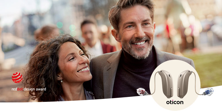 Two blurred individuals pose outdoors in a social setting. Featured in the foreground are Oticon hearing aids, highlighted alongside the Red Dot Design Award logo, symbolizing cutting-edge hearing aid technology.
