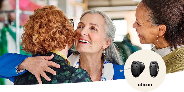 Two individuals embrace warmly in what appears to be a social setting, with another person nearby. An inset highlights Oticon hearing aids, suggesting enhanced hearing support through advanced technology.
