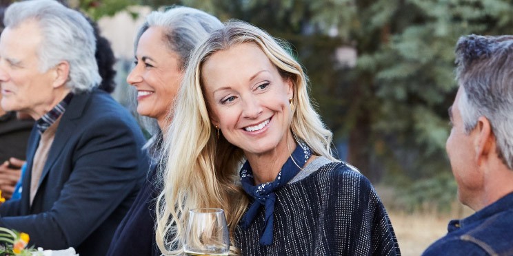 Woman enjoying a conversation outdoors while holding a drink, surrounded by others at a table.