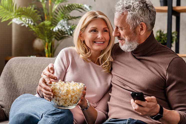 Couple sitting on a sofa eating popcorn and watching television, with a remote control in hand, in a cozy home setting.