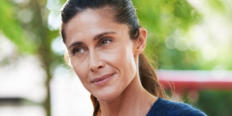 Woman wearing a hearing aid outdoors with blurred green foliage in the background.