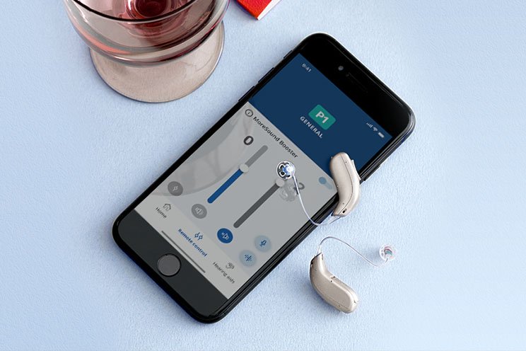 Mobile phone displaying hearing aid app interface alongside two modern hearing aids.