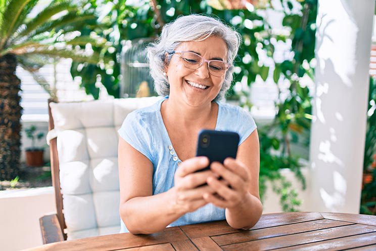 Senior woman with short gray hair using a smartphone while sitting outdoors at a wooden table surrounded by greenery.
