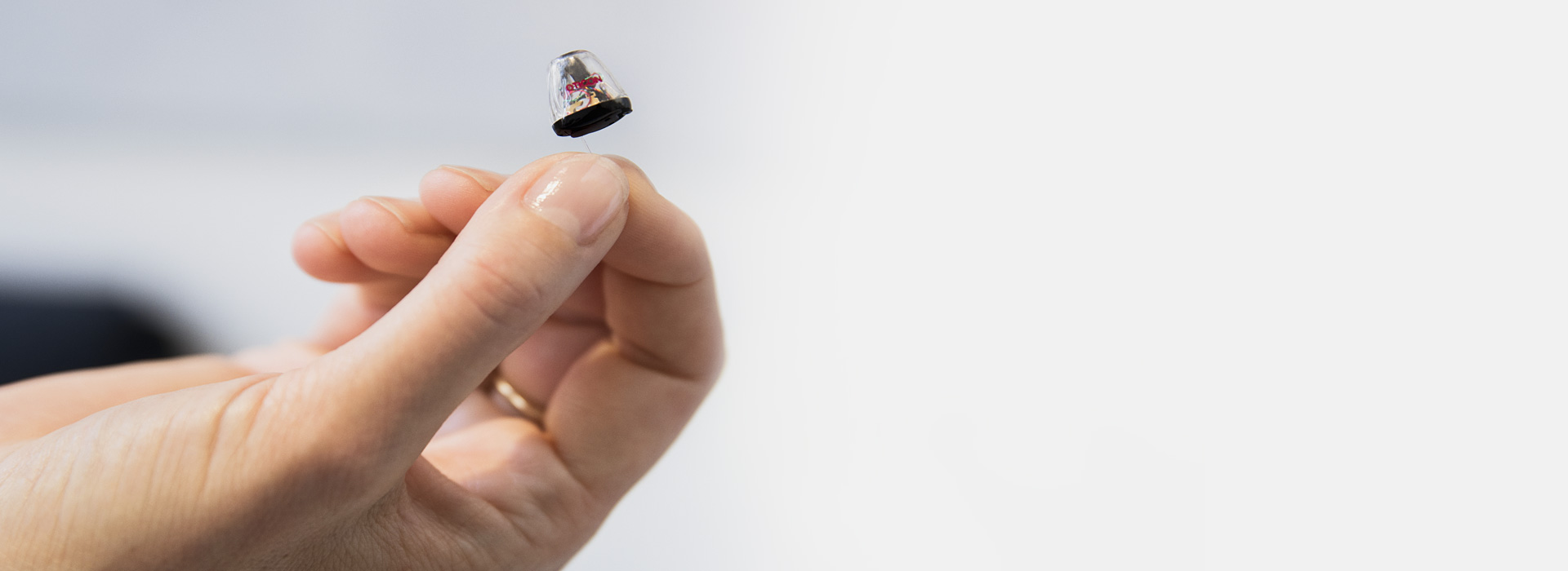 A hand holds a discreet hearing aid with a transparent top between the thumb and index finger, set against a clean white background.