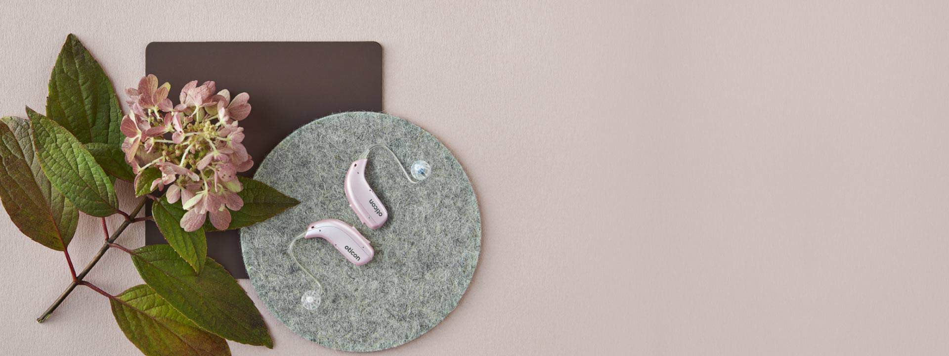 Pink Oticon hearing aids resting on a textured gray circle, accompanied by pink hydrangea flowers and green leaves, set against a soft beige background.