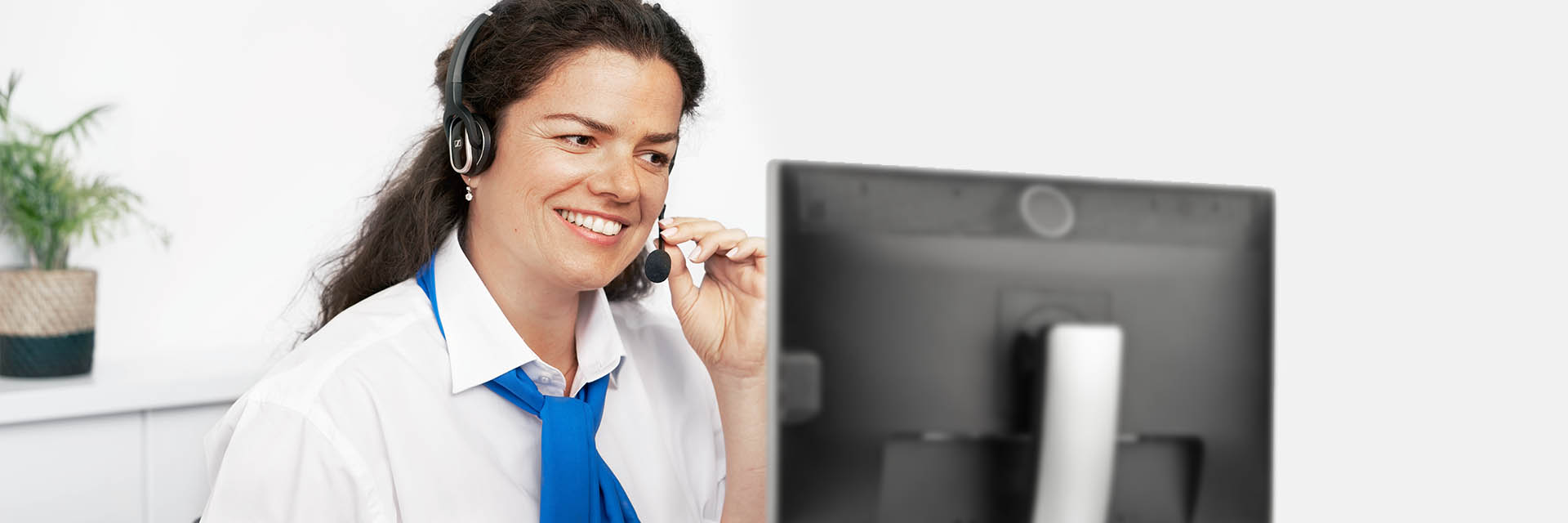 A customer service representative wearing a headset speaks into a microphone, sitting in front of a computer monitor in a bright office with a potted plant on the desk.