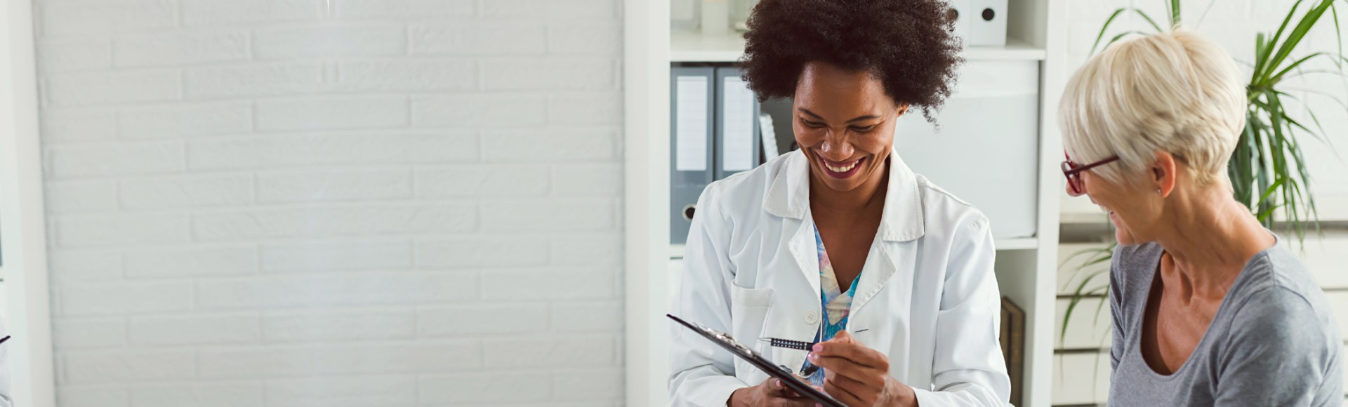A smiling doctor in a white coat writes notes on a clipboard while interacting with an elderly woman in a brightly lit office featuring shelves, binders, and indoor plants.