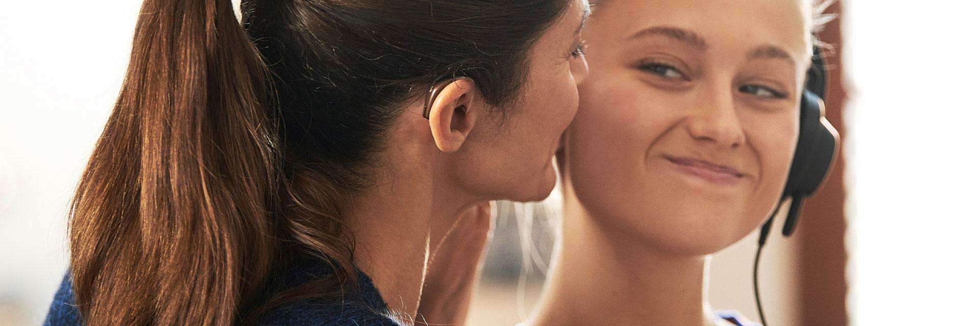 Woman wearing a hearing aid smiling warmly at a young girl using headphones.