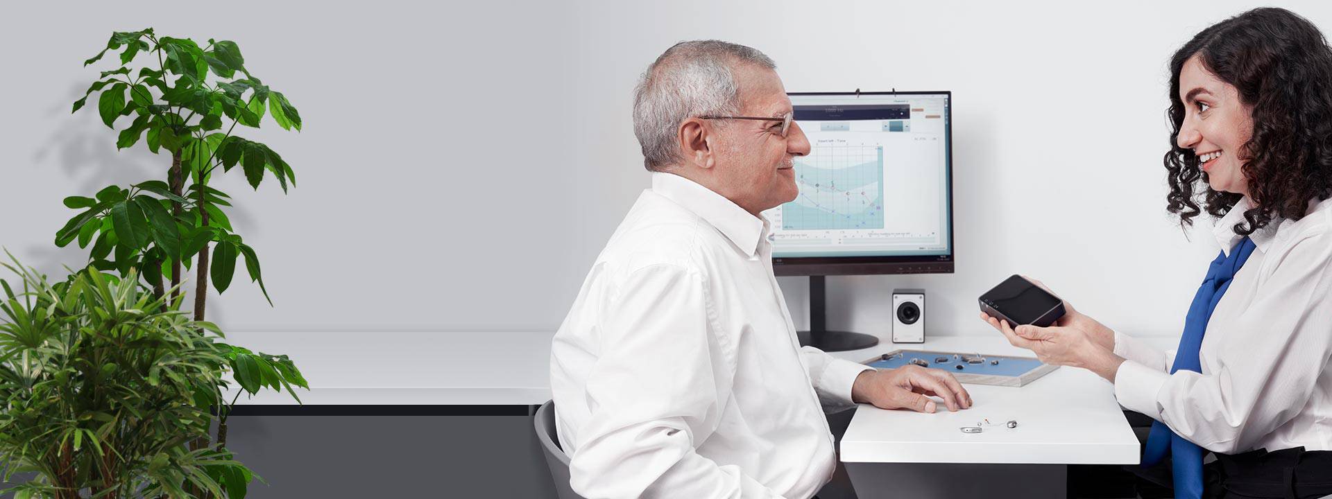 Man discussing hearing aid options with an audiologist, with accessories and an audiogram display visible on a desk.