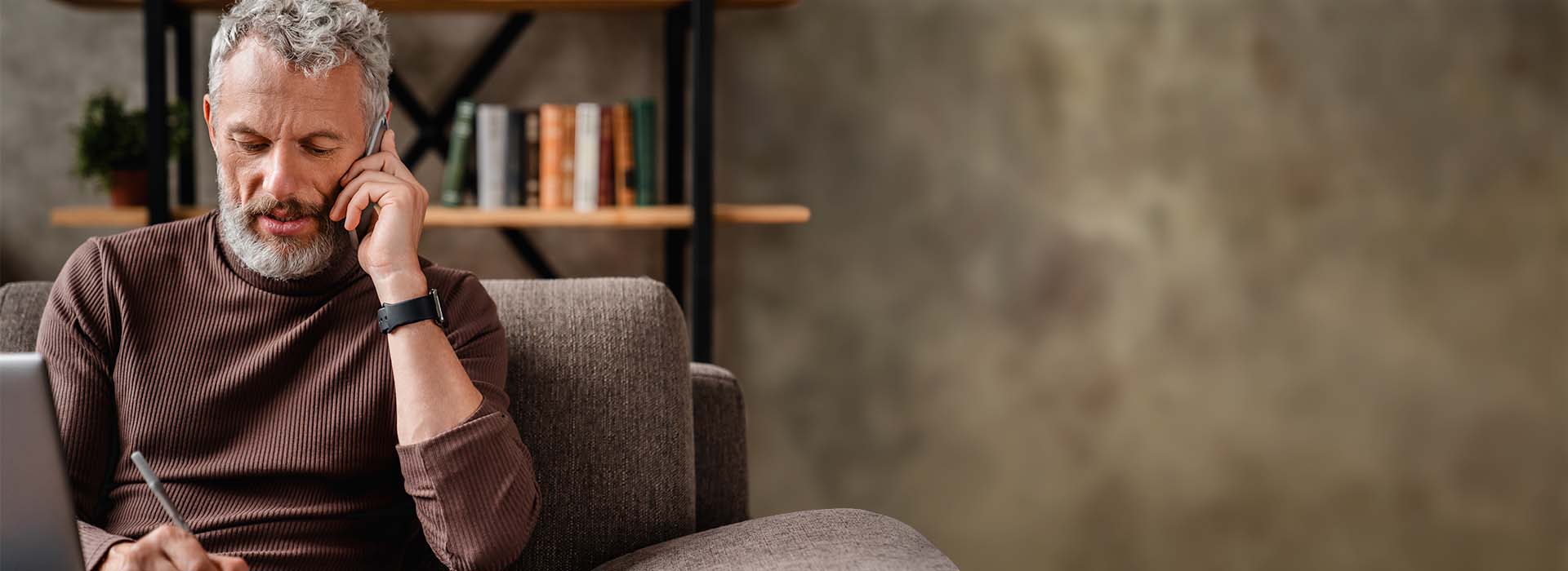 Man wearing a hearing aid talking on the phone and taking notes while seated on a comfortable sofa.