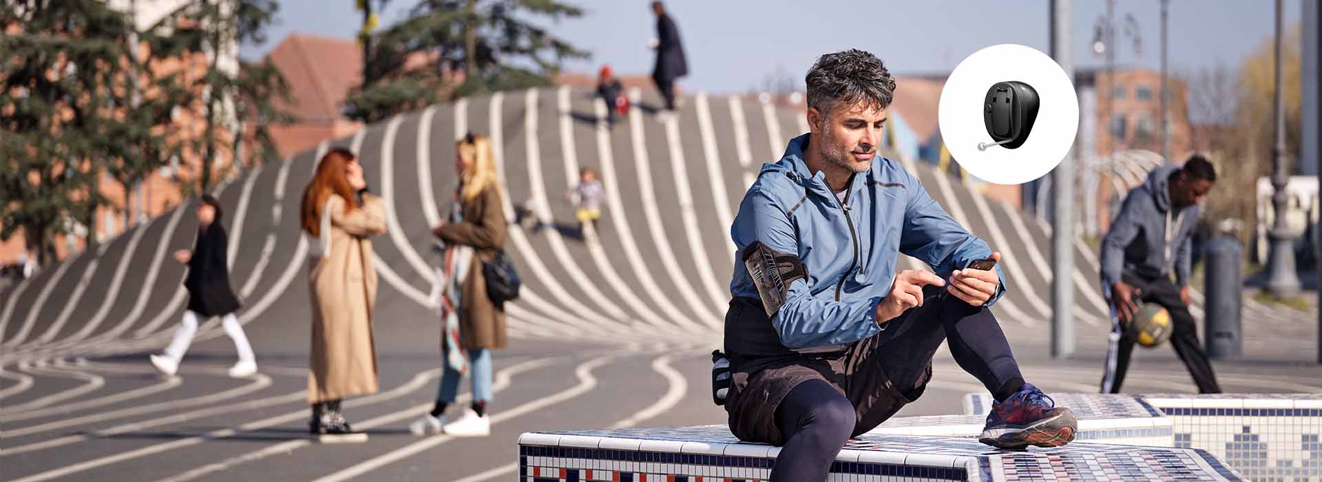 A person sits on a tiled bench outdoors, focused on their phone. Nearby, people interact and move across a striped, undulating surface in an urban setting.