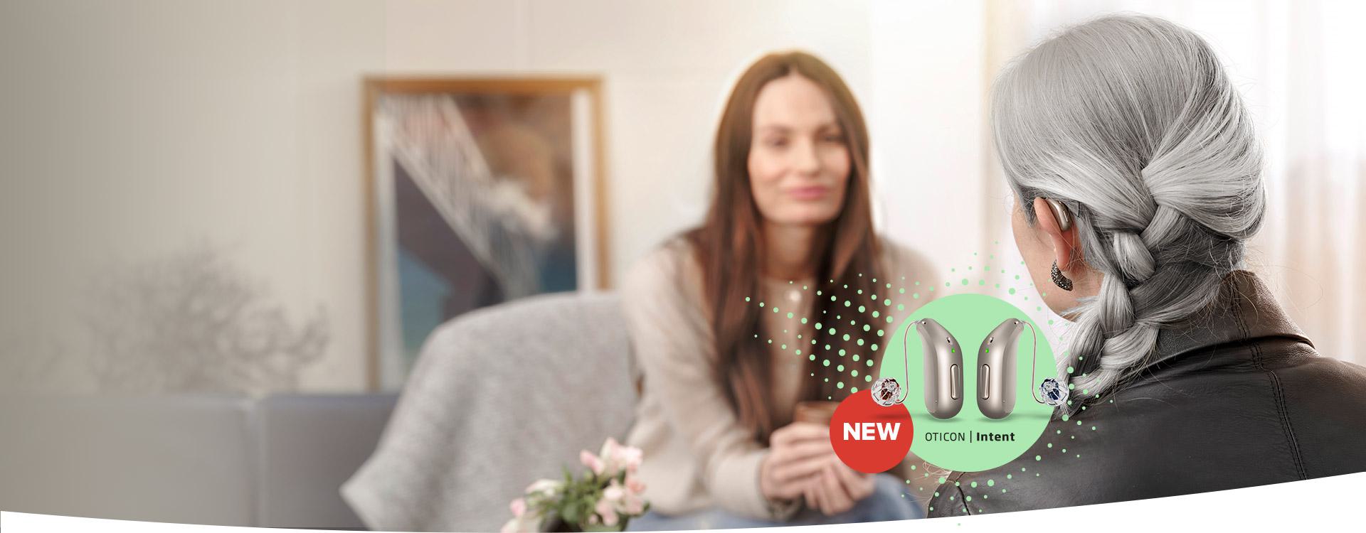 An elderly woman with grey braided hair wearing a hearing aid engages in conversation with a younger woman in a warmly decorated living room, alongside a promotional display of the new Oticon Intent hearing aids.