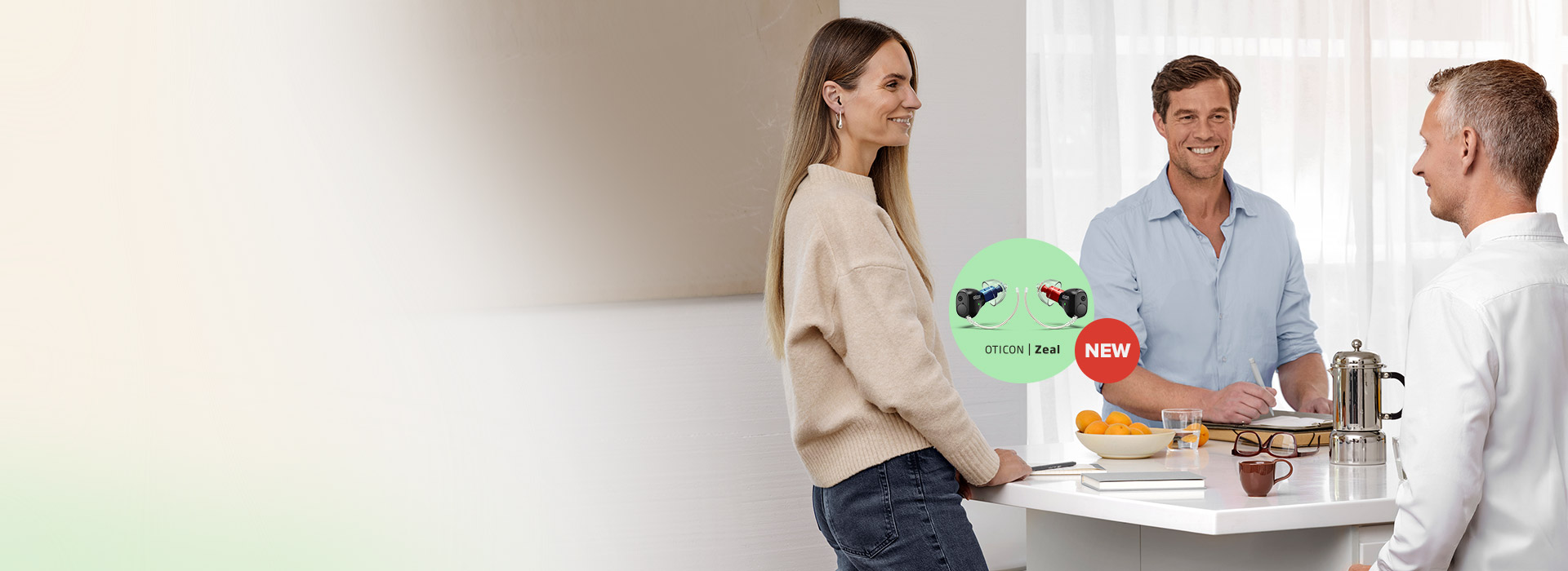 Three individuals converse around a white counter in a brightly lit kitchen. A green bubble highlights 'Oticon Zeal hearing aids', labeled 'NEW,' emphasizing advanced hearing care products amid a casual, friendly setting.