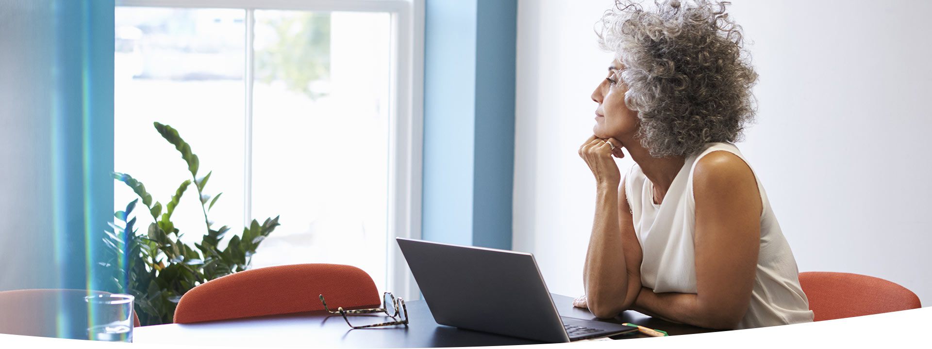 A woman with curly gray hair rests her chin on her hand while sitting at a table with a laptop, eyeglasses, and a glass of water; sunlight illuminates the room with a plant near the window.