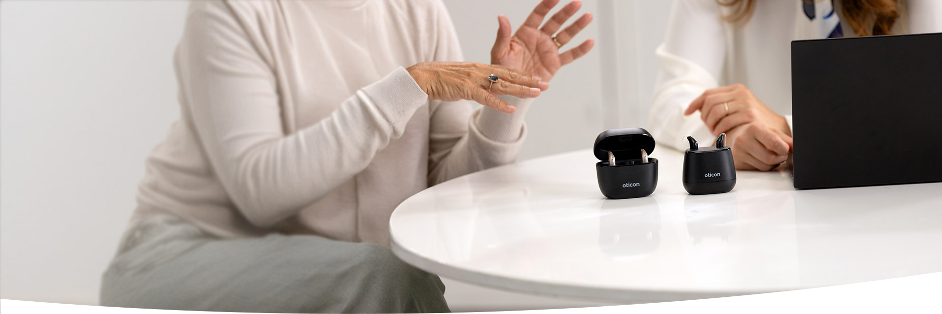 Two black Oticon hearing aid cases, one open displaying devices, are placed on a white table. Nearby, hands gesture in conversation next to a laptop in a professional hearing clinic.