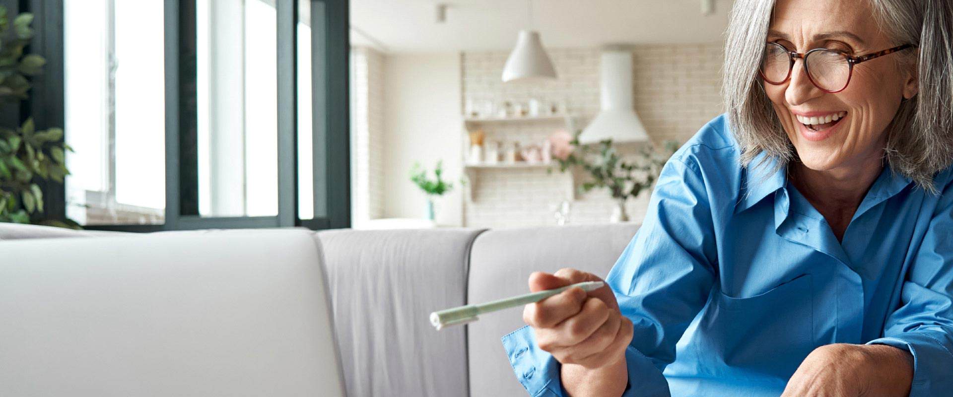 Woman with gray hair wearing a blue shirt and holding a hearing aid in a bright, modern living room.