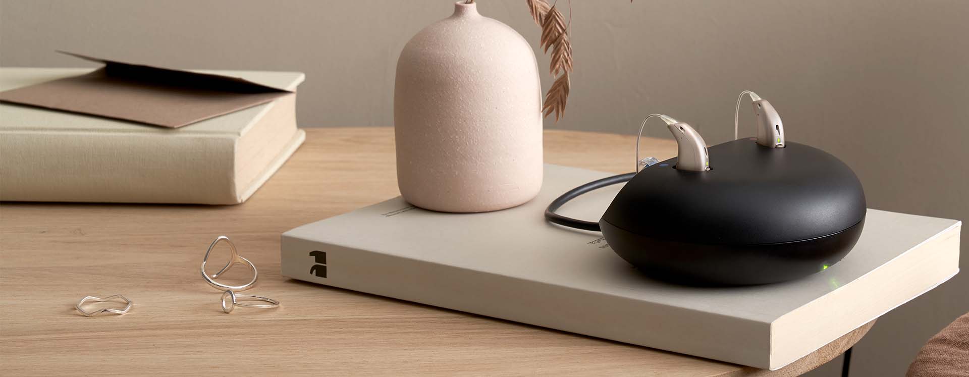 Hearing aids placed on a sleek black charging case on a wooden table alongside a decorative vase, books, and minimalist rings.