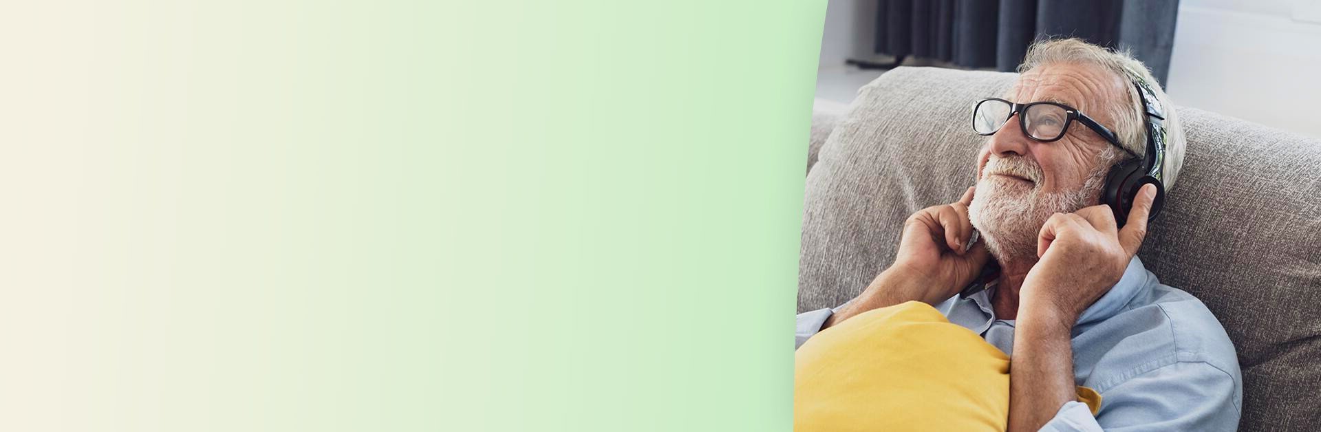 Man wearing glasses and headphones, smiling while lying on a couch with a yellow pillow; light green gradient background on the left, and dark curtains in the background.
