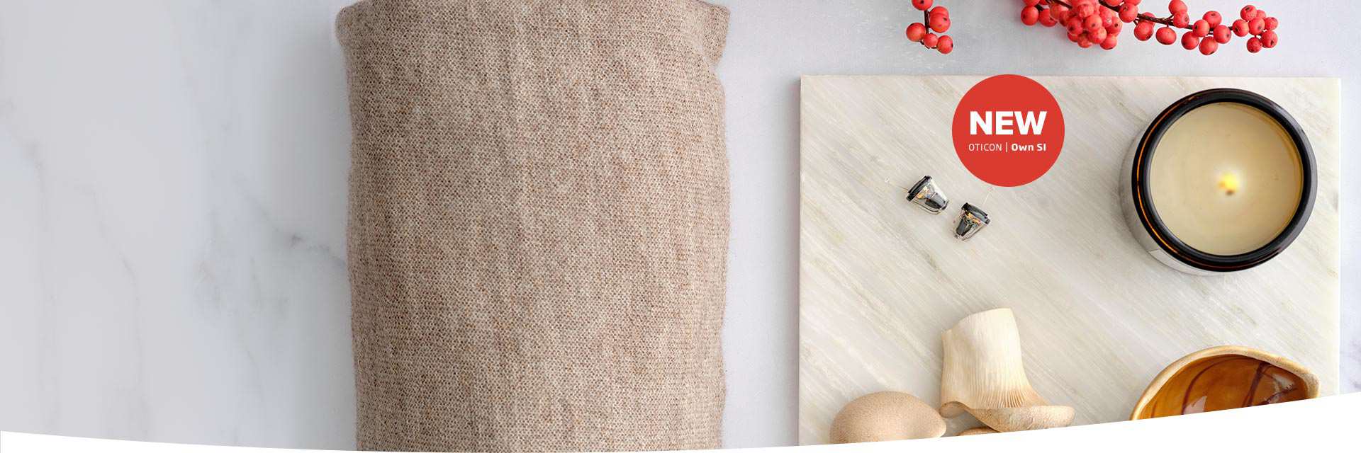Two Oticon Own SI hearing aids rest on a light marble surface near a lit candle, decorative red berries, and a beige textured fabric. Text reads: ’NEW OTICON | Own SI.’