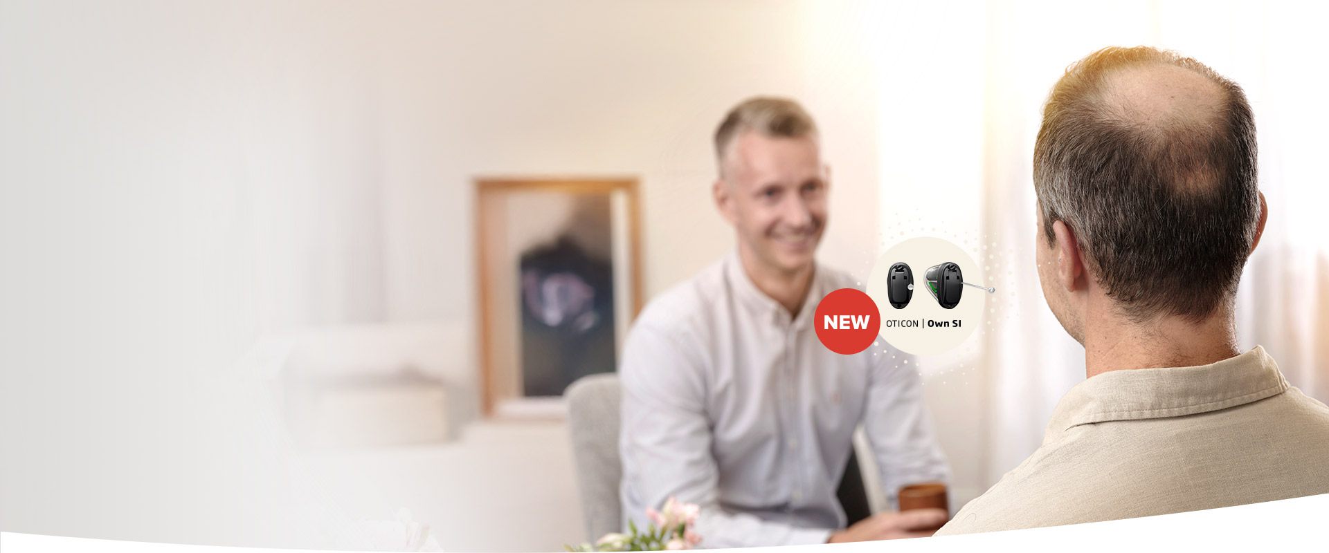 A balding man faces an audiologist seated opposite him during a consultation in a softly lit room. A promotional ’NEW Oticon Own S1’ hearing aid graphic appears on the right.