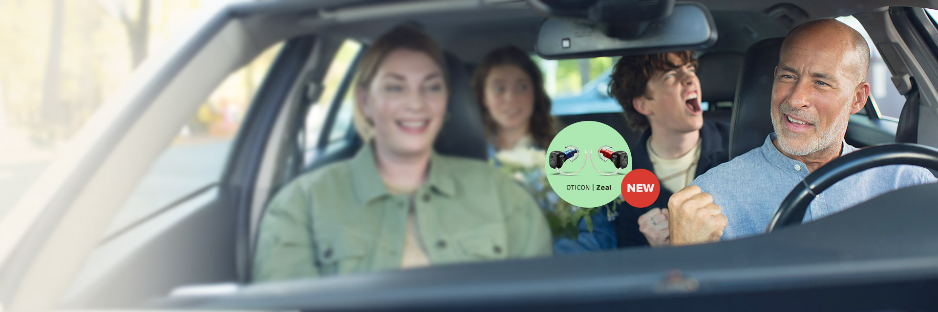 A smiling man drives a car with three cheerful passengers. An overlay promotes 'Oticon Zeal' hearing aids, showing two devices (red and blue) alongside a 'NEW' badge.