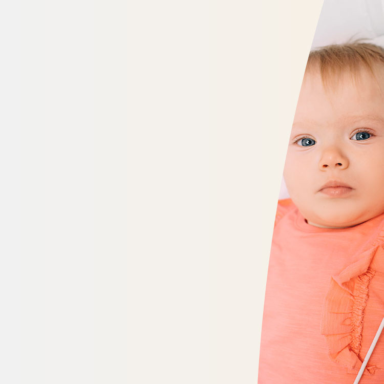 A baby with blue eyes and light hair looks forward, wearing a coral outfit accented with ruffles. The asymmetrical composition places the baby in the right section, surrounded by an off-white background.
