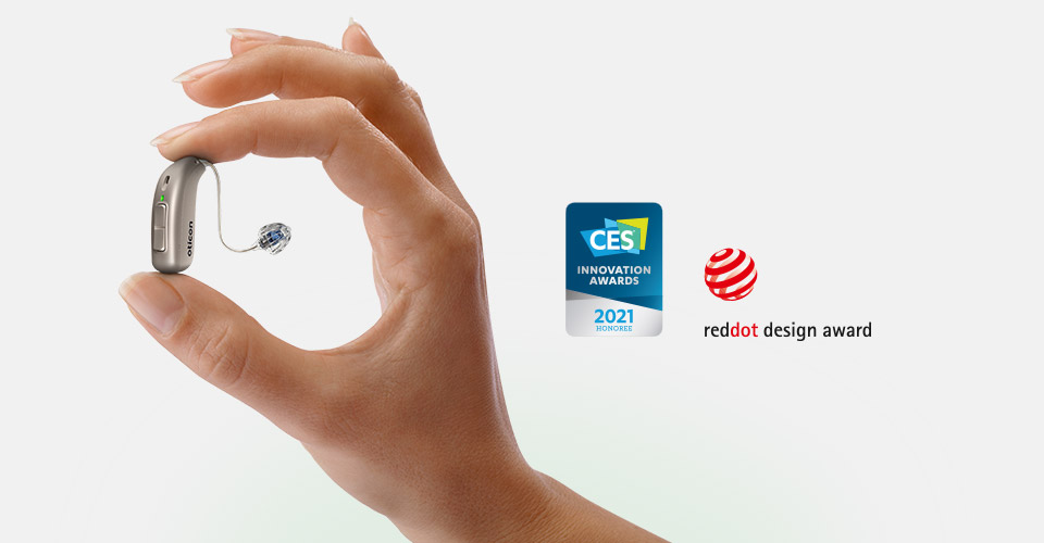 A hand holds an Oticon hearing aid between thumb and forefinger, showcasing its compact design. To the right, awards are displayed: ’CES Innovation Awards 2021 Honoree’ and ’Red Dot Design Award.’