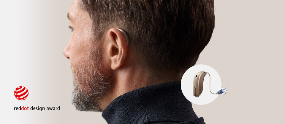 A lightweight, discreet hearing aid rests behind a man’s ear, showcasing advanced design and functionality. The Red Dot Design Award logo is displayed, emphasizing innovation. Inset shows a close-up of the hearing aid.
