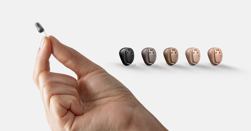 A hand holding a small hearing aid device, with five hearing aids in black, brown, and beige colors arranged in a row on the right side against a white background.