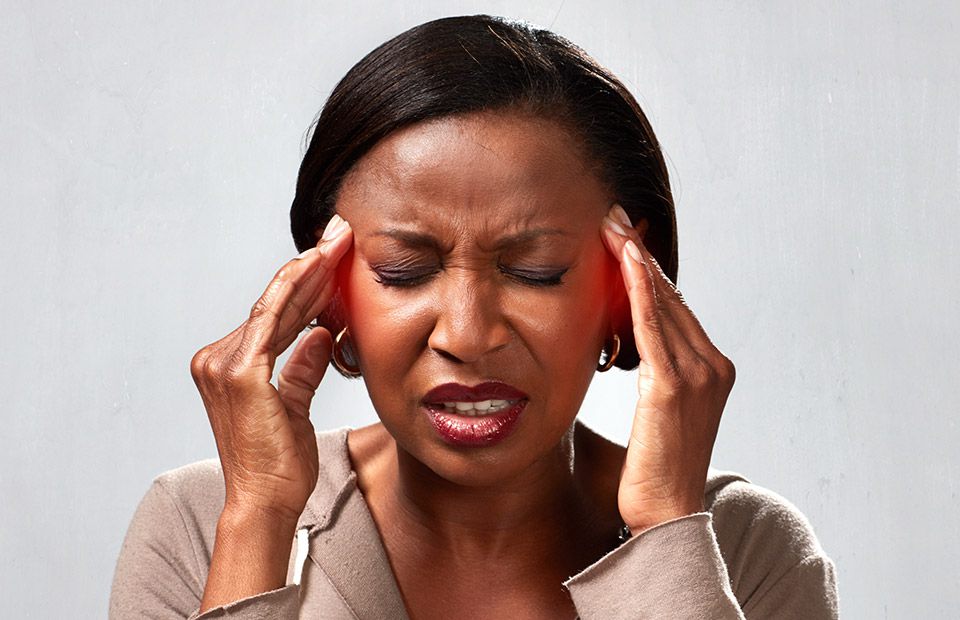 Woman with closed eyes pressing her temples with both hands, possibly experiencing discomfort, stress, or a headache, against a neutral grey background.