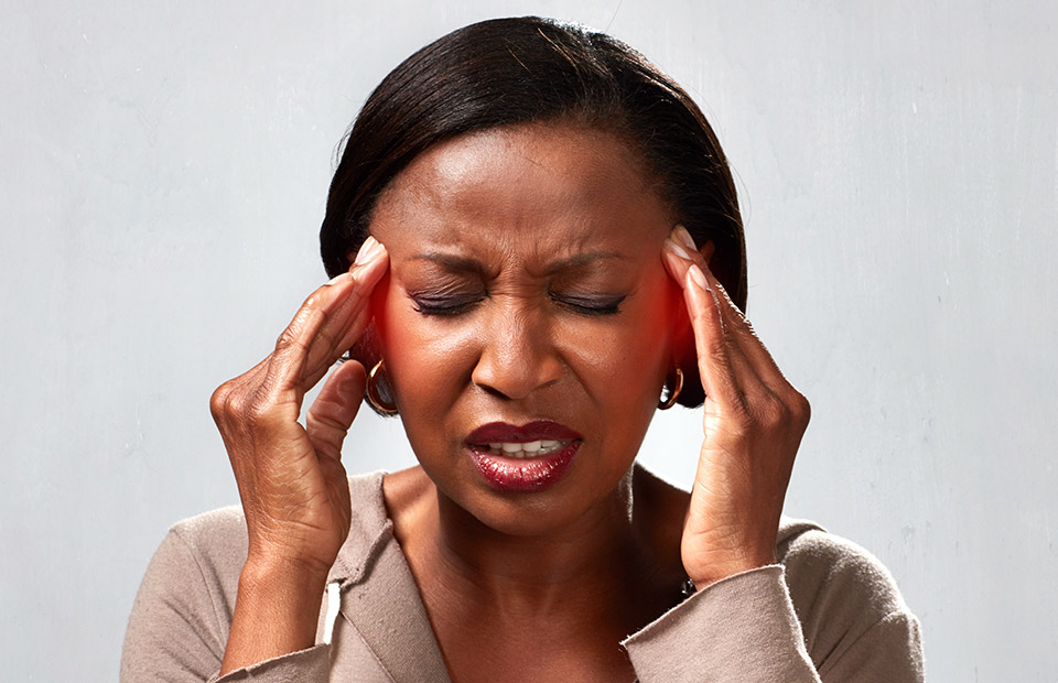 Woman pressing her temples with both hands, eyes closed, on a grey background, appears to be experiencing a headache or stress.