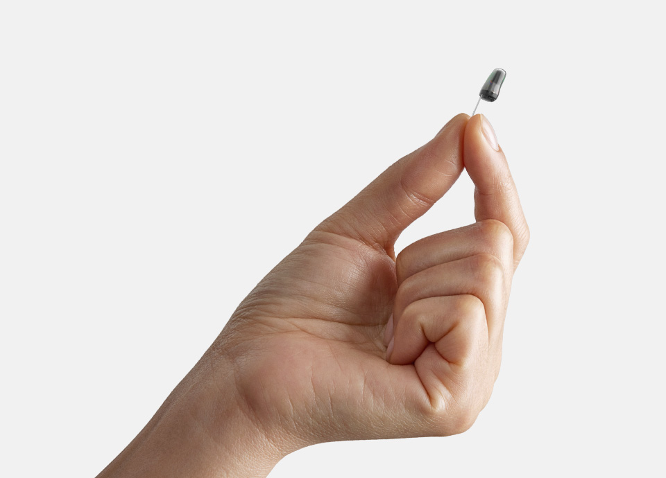 A hand holds a small, gray hearing aid between the thumb and forefinger against a plain, light background.