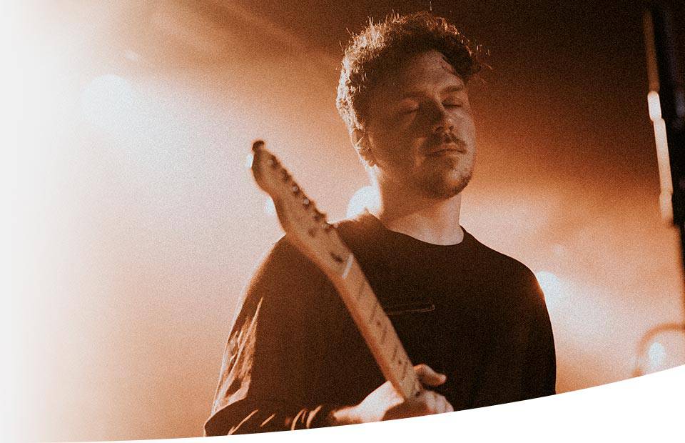 A guitarist performs under warm, focused stage lighting in a dim concert atmosphere, holding an electric guitar with a blurred background to highlight the subject.