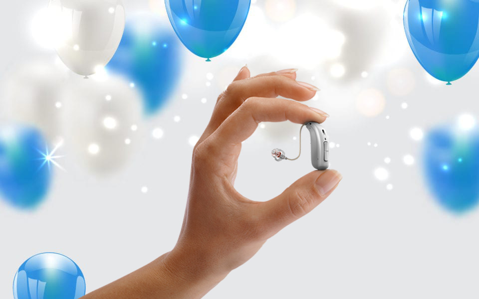 A hand holds a small, modern hearing aid device against a background of floating blue and white balloons with soft glowing lights.