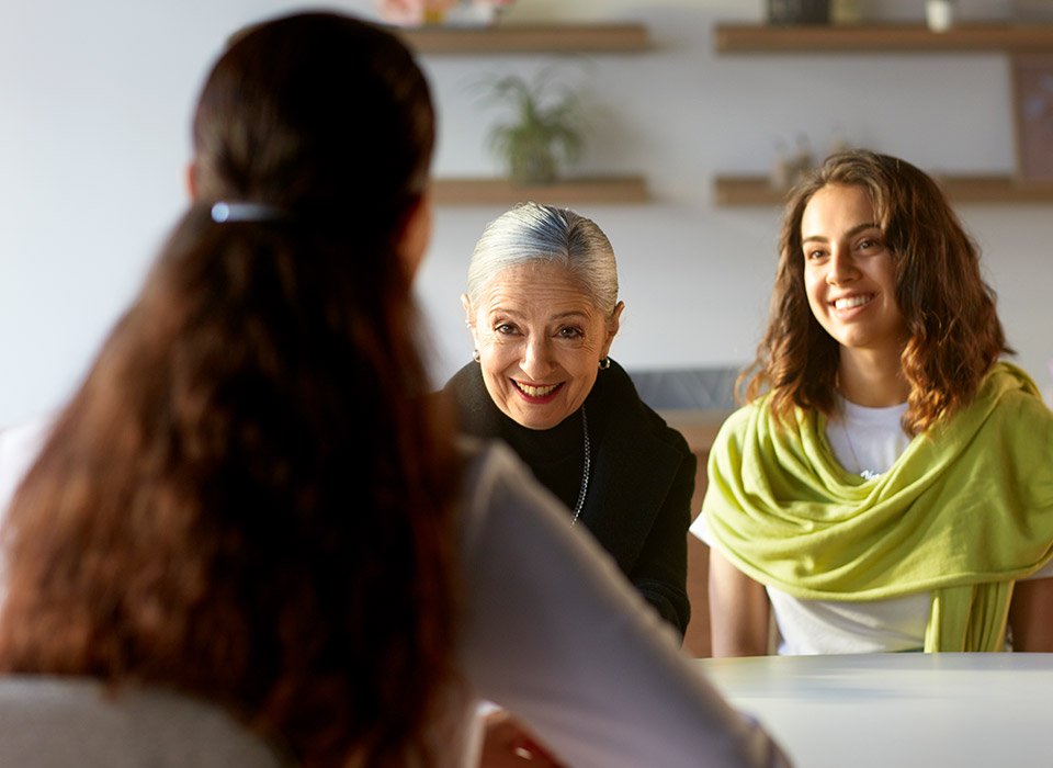 A woman with long brown hair, viewed from behind, appears to be in a discussion with two women seated across a table in a bright, modern room with shelves in the background.