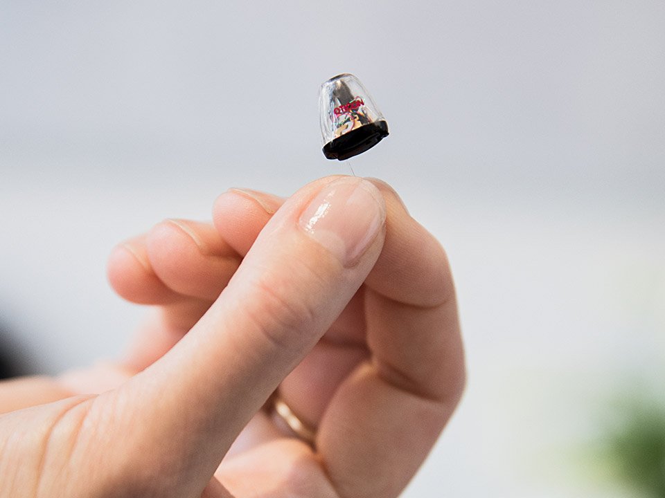 Hand holding a discreet in-ear hearing aid against a blurred background.