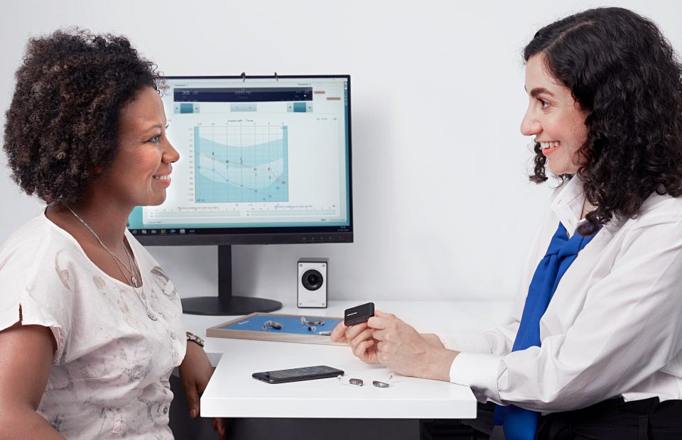 Woman consulting with a hearing care professional while trying on hearing aids, with a computer displaying an audiogram in the background.
