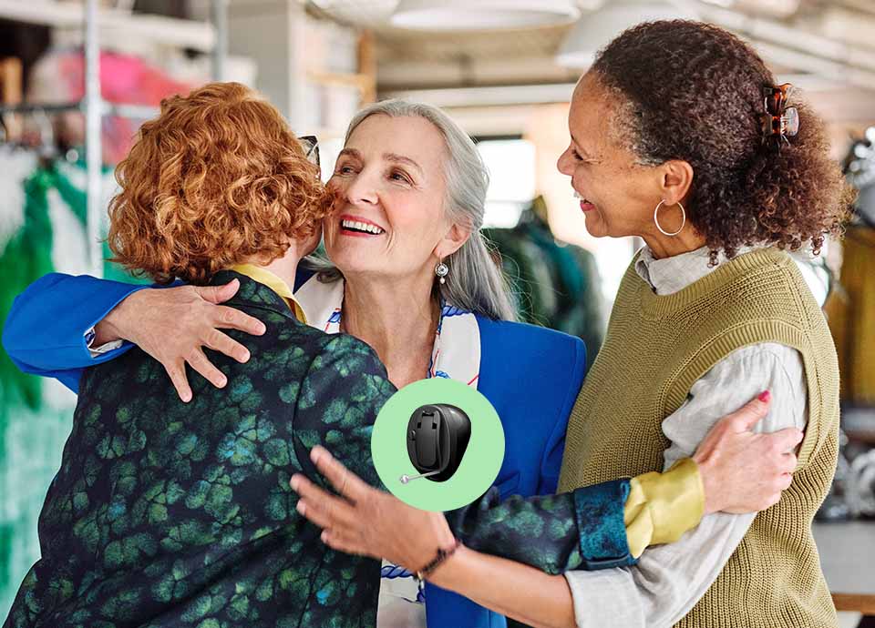 Three women embrace warmly in a brightly lit, clothing-filled workspace. A featured black hearing aid, shown on a circular green overlay, highlights hearing care solutions from HearingLife.