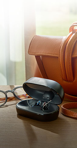 A pair of hearing aids rests in a sleek black charging case on a wooden table, surrounded by eyeglasses and an orange leather bag near a sunlit window.