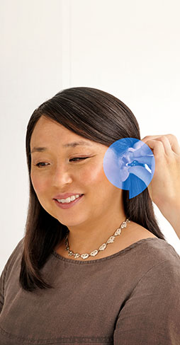 A smiling woman wearing a brown top sits in a hearing clinic. An audiologist places a hearing aid in her ear, highlighted by a blue magnified overlay.