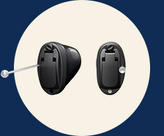Two black in-ear hearing aids are set against a circular beige background with a navy blue border. One hearing aid has a small tool inserted, possibly for adjustment or cleaning.