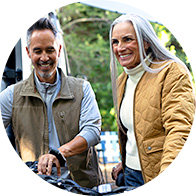 A man and woman, wearing casual outdoor jackets, examine a bicycle in an outdoor setting surrounded by trees. Their facial features are blurred for privacy. No text or identifiable hearing aids are visible.