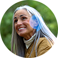 A silver-haired individual wearing a beige jacket and white turtleneck is outdoors in a green, nature-filled environment. A discreet hearing aid is visible behind their ear, enhancing hearing ability.