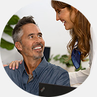A man sits in a hearing clinic while a professional, possibly an audiologist, places a reassuring hand on his shoulder. A laptop and leafy plants are visible in the background.