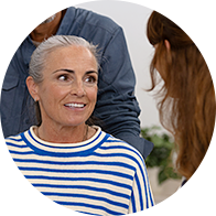 An older individual wearing a blue-and-white striped shirt sits while interacting with a hearing care professional in a clinical setting decorated with plants, highlighting a hearing consultation at a clinic. No text visible.