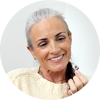 A senior individual with gray hair is holding a discreet hearing aid in their hand. The setting appears to be a bright, clean space, likely a hearing clinic. No text is visible.