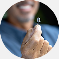 A hand holds a discreet hearing aid between fingers, with a smiling individual blurred in the background, suggesting a positive experience at a hearing clinic like HearingLife. No visible text.