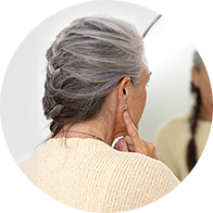 A woman with braided gray hair adjusts a hearing aid behind her ear while standing in front of a mirror in a well-lit setting, wearing a beige sweater. No text visible.