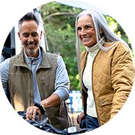 A man and woman, wearing casual outdoor jackets, examine a bicycle in an outdoor setting surrounded by trees. Their facial features are blurred for privacy. No text or identifiable hearing aids are visible.