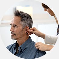 An audiologist fits a hearing aid on an adult man’s ear in a brightly lit hearing clinic.